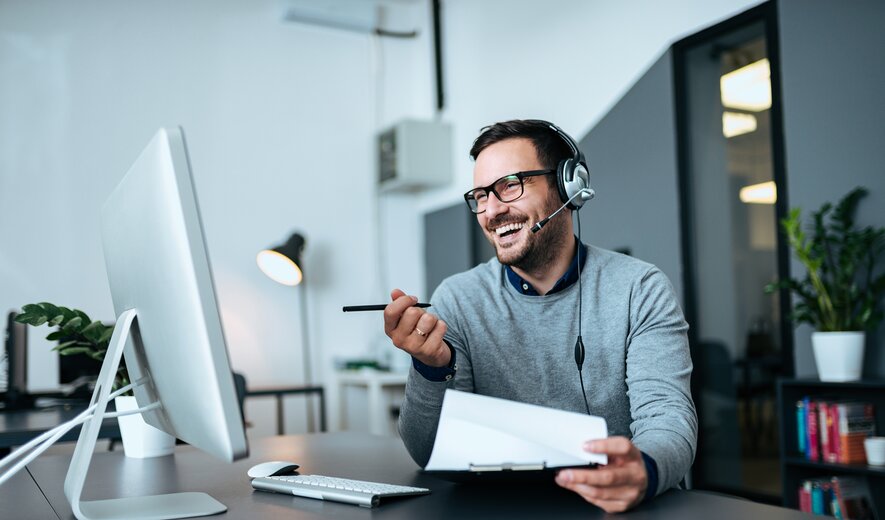 Mitarbeiter im Büro führt ein fröhliches Online-Meeting mit Headset am Computer durch.